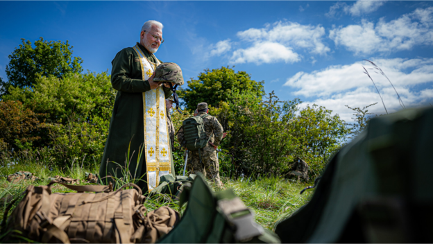 Ein ukrainischer Militärseelsorger hält den Helm eines Soldaten auf einem Truppenübungsplatz. Die Kirche kann bei existenziellen Fragen auch außerhalb von Krieg und Katastrophen Antworten bieten.
