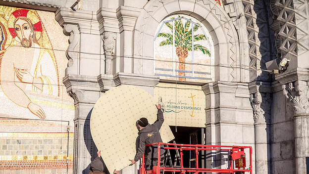 Arbeiter halten eine Abdeckung, die zur Verhüllung des Mosaiks am Eingang der Rosenkranz-Basilika angebracht wird, am 31. März 2025 in Lourdes (Frankreich). Das Mosaik ist ein Kunstwerk von Priester und Künstler Marko Ivan Rupnik, der unter Verdacht steht, sexuellen Missbrauch begangen zu haben.